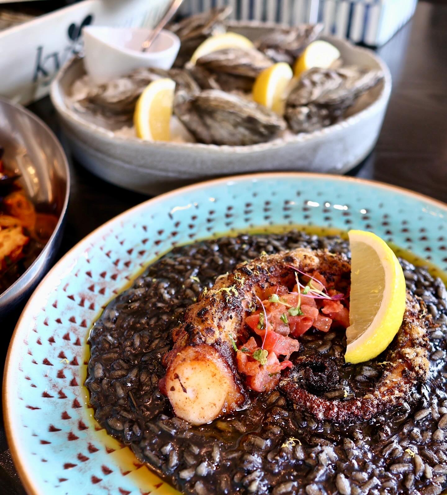 Deep black squid ink risotto served with a large grilled octopus tentacle, diced tomato garnish, and a lemon wedge, with fresh oysters in the background.